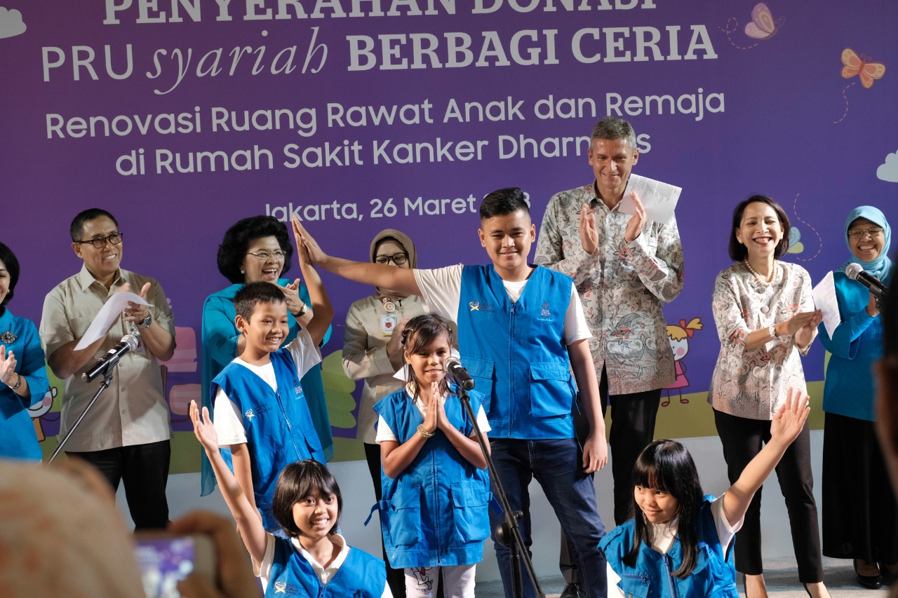 A group of children and adults stand on a stage during a charity event. The children in the front wear matching blue vests while speaking or performing with microphones. Behind them, adults applaud and smile against a purple backdrop with butterflies and clouds. Indonesian text on the backdrop mentions a donation handover, renovation of pediatric and teen wards at a cancer hospital, and the date Jakarta, 26 Maret 2017.