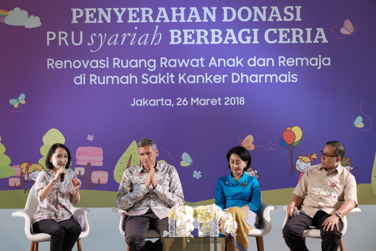 Four adults sit on a stage during a formal donation event, speaking into microphones in front of a purple backdrop. The Indonesian text announces a donation handover and renovation of pediatric and adolescent wards at Rumah Sakit Kanker Dharmais. The backdrop includes the date Jakarta, 26 Maret 2018 and playful illustrations of clouds, butterflies, and balloons, creating a cheerful atmosphere. A small table with white flowers is placed between the speakers, emphasizing the ceremonial nature of the occasion.