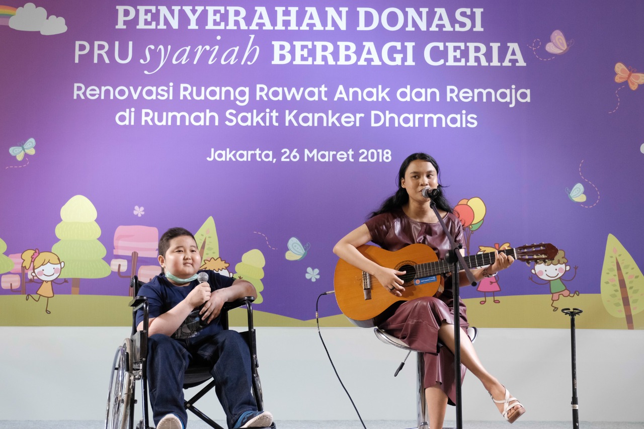 A young woman plays an acoustic guitar and sings on stage beside a boy in a wheelchair during a charity donation event. The backdrop text in Indonesian mentions the handover of donations for renovating pediatric and teen wards at a cancer hospital. The event takes place in Jakarta on 26 Maret 2018, with a colorful illustrated background featuring children, clouds, and butterflies. The atmosphere is warm and uplifting, highlighting support and joy for young patients.
