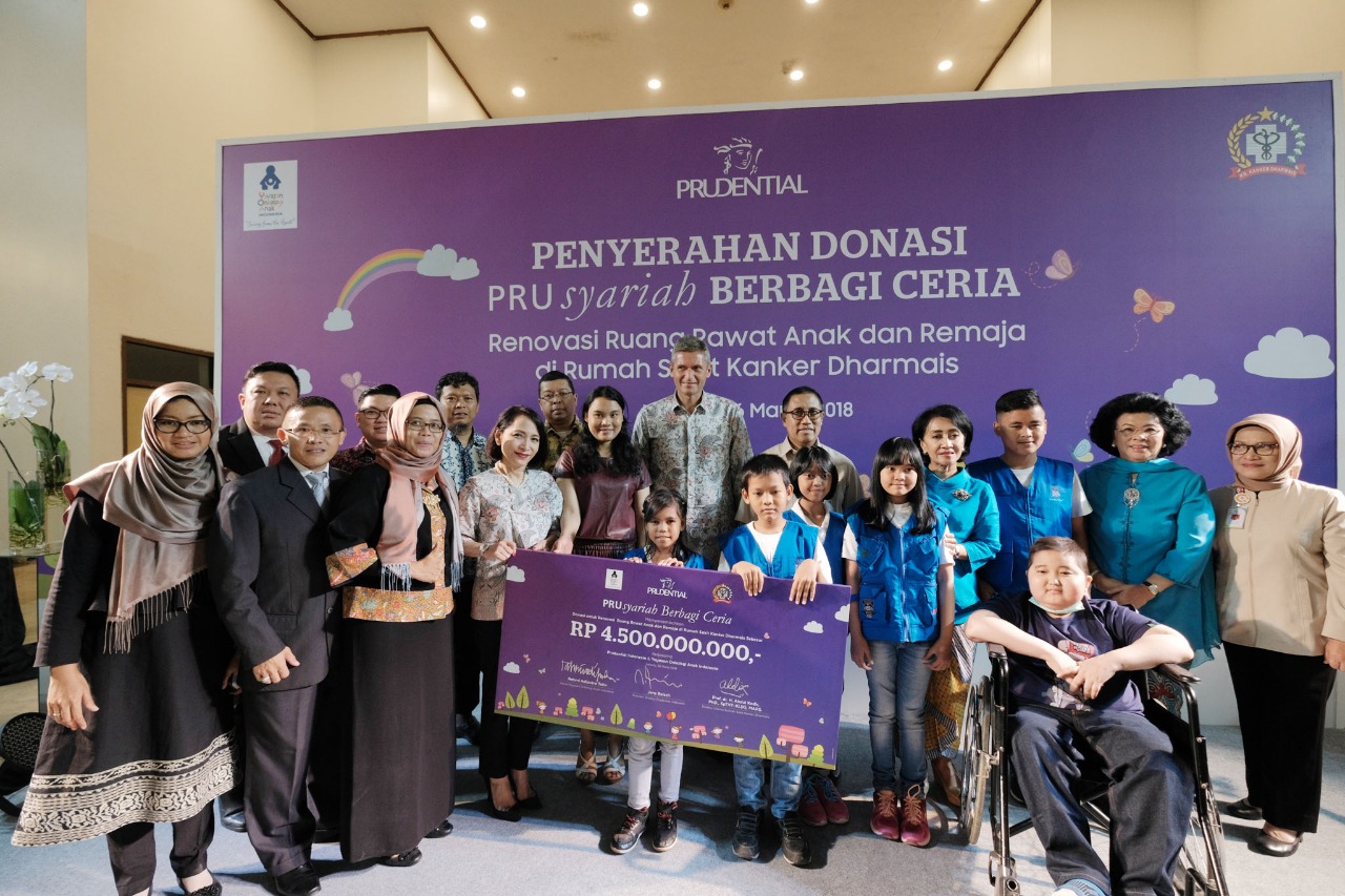 A group of adults and children pose together at a charity donation event, holding a large ceremonial check. The backdrop text in Indonesian references a donation handover and renovation of pediatric and adolescent wards at a cancer hospital. The check prominently displays the amount RP 4.500.000.000,00 and colorful illustrations of a cityscape and children. Participants include healthcare staff, organization representatives, and young patients, one seated in a wheelchair, all gathered in a bright indoor venue. The atmosphere is warm and celebratory, emphasizing community support and social responsibility.