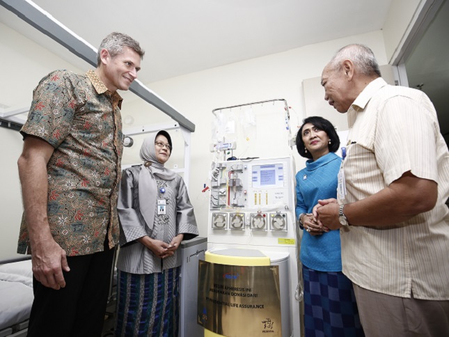A group of adults stands around a large medical device in a clinical room. The machine features multiple tubes, dials, and a digital display panel, suggesting specialized hospital equipment. Two women in traditional attire and two men in casual shirts appear to be discussing or inspecting the device. The setting looks like a hospital or healthcare facility, with white walls and a bed partially visible in the background.