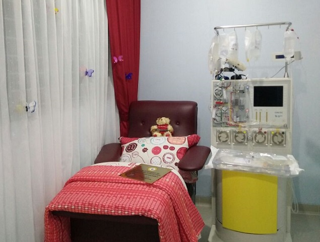 A comfortable medical treatment chair is set up beside a dialysis or infusion machine in a small hospital room. The chair is covered with a red checkered blanket and patterned pillow, and a teddy bear rests on the seat to create a comforting atmosphere. The medical machine stands on a yellow base with tubes and fluid bags hanging above it. Sheer white curtains with small colorful butterfly decorations soften the clinical setting and add a touch of warmth.