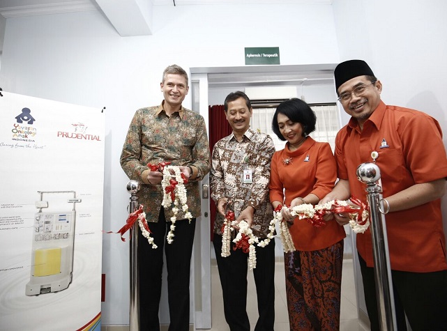 Four adults stand together at a ribbon cutting ceremony in an indoor medical or clinical facility. They hold floral garlands and red ribbons between two stanchions, indicating the opening of a new room or service. A poster on the left shows a medical device illustration and the word Prudential, suggesting a healthcare-related initiative. The participants wear patterned shirts and uniforms, and a green sign is visible above the doorway in the background.