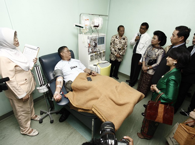 A patient lies on a recliner chair undergoing a medical blood procedure in a clinical room. A healthcare professional stands nearby holding a clipboard while monitoring the patient and equipment. Several formally dressed visitors stand around, observing the procedure and listening attentively. Medical devices, tubing, and a blanket covering the patient emphasize the hospital environment and healthcare context.