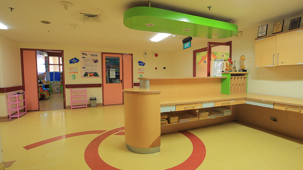 A brightly colored reception desk stands in the center of a pediatric hospital or clinic corridor. The space features pastel yellow floors with red circular patterns and light orange walls. Pink door frames and furniture, along with a green overhead structure, create a playful, child-friendly environment. Posters and signs are visible on the walls, and an EXIT sign hangs above one of the doors.