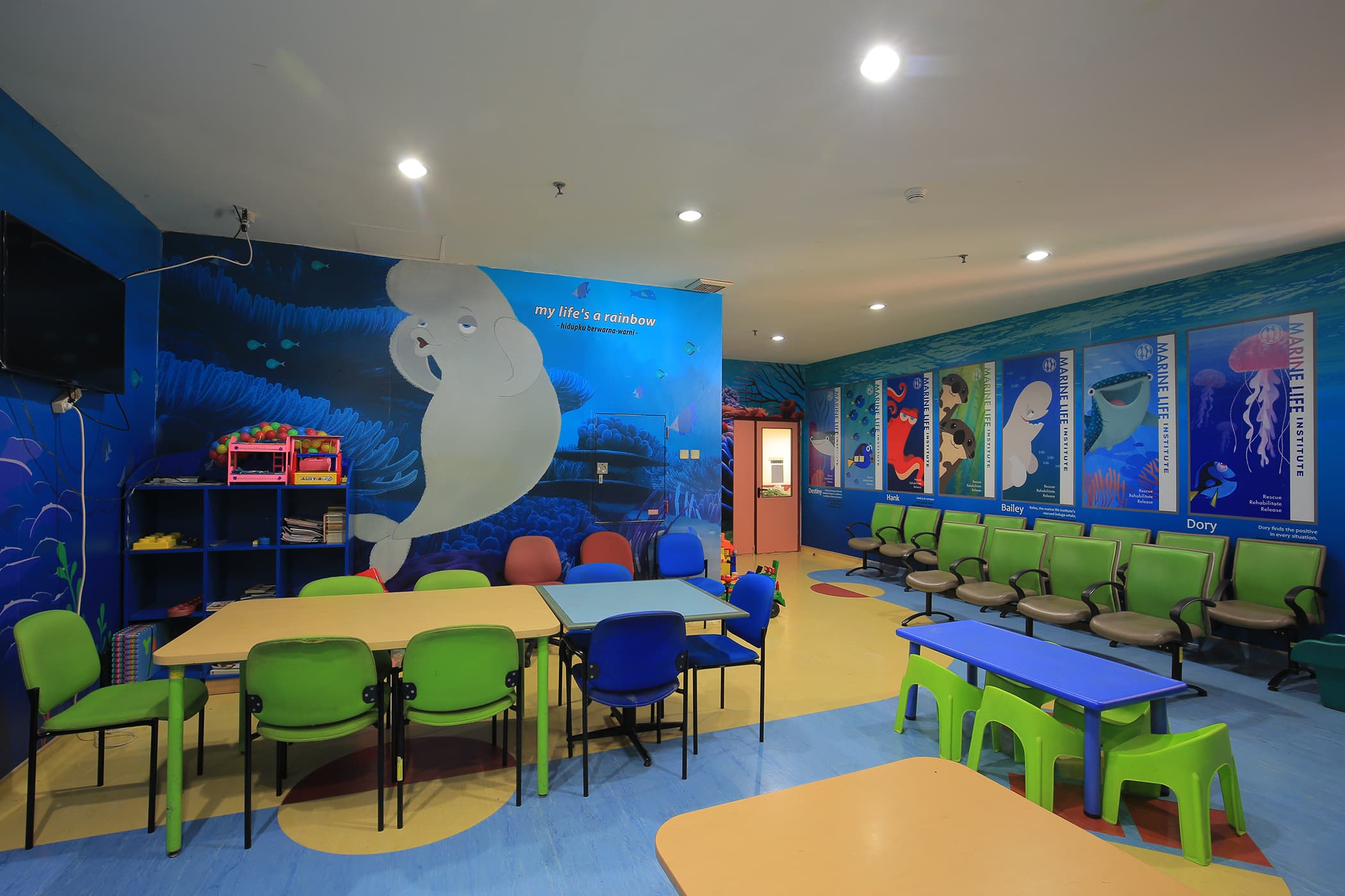 Brightly colored children's classroom decorated with an underwater ocean theme. Walls feature large illustrations of sea animals, including a manatee and jellyfish, with educational posters. Small tables and chairs in green, blue, and yellow are arranged for group activities, with a row of cushioned seats along one wall. Toys and supplies are neatly stored on shelves, creating an inviting learning environment.