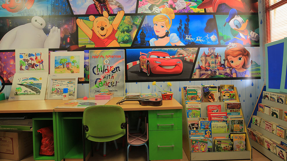 A bright children's reading and activity corner is decorated with large cartoon character murals above a desk and book display. The room features a green chair, art supplies, and children's drawings arranged along the work surface. A poster reading "Children with Cancer" is prominently displayed, suggesting a supportive or hospital environment. A rack filled with colorful children's books stands to the right, adding to the playful and welcoming atmosphere.