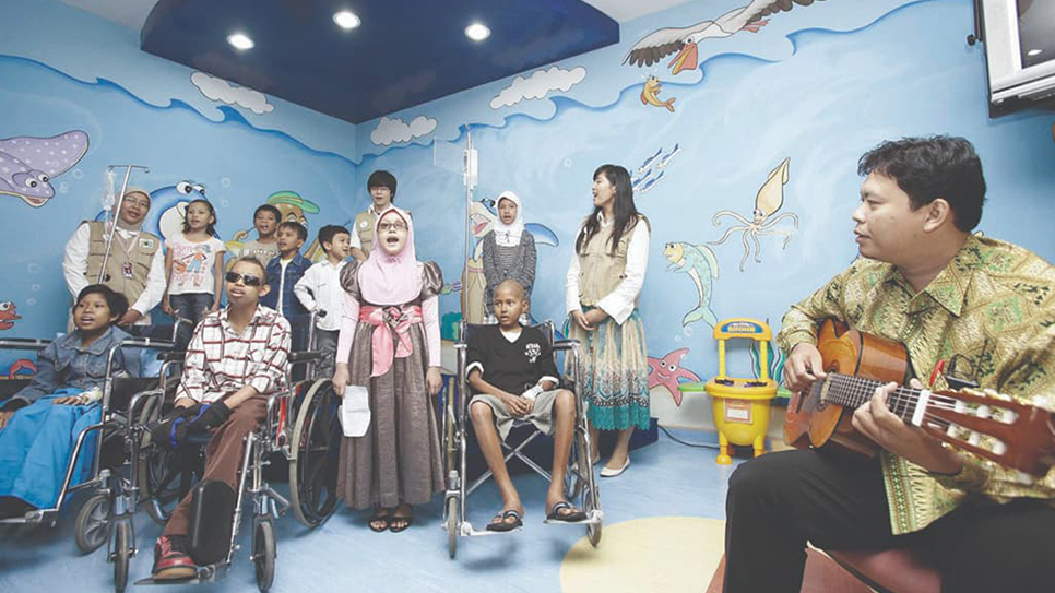 A group of children, some in wheelchairs and with IV stands, gather in a brightly decorated hospital room. A man sits to the side playing an acoustic guitar while the children listen and sing along. The walls feature colorful ocean-themed murals with fish and sea creatures, creating a cheerful environment. Several adults stand nearby, offering support and encouragement to the young patients.