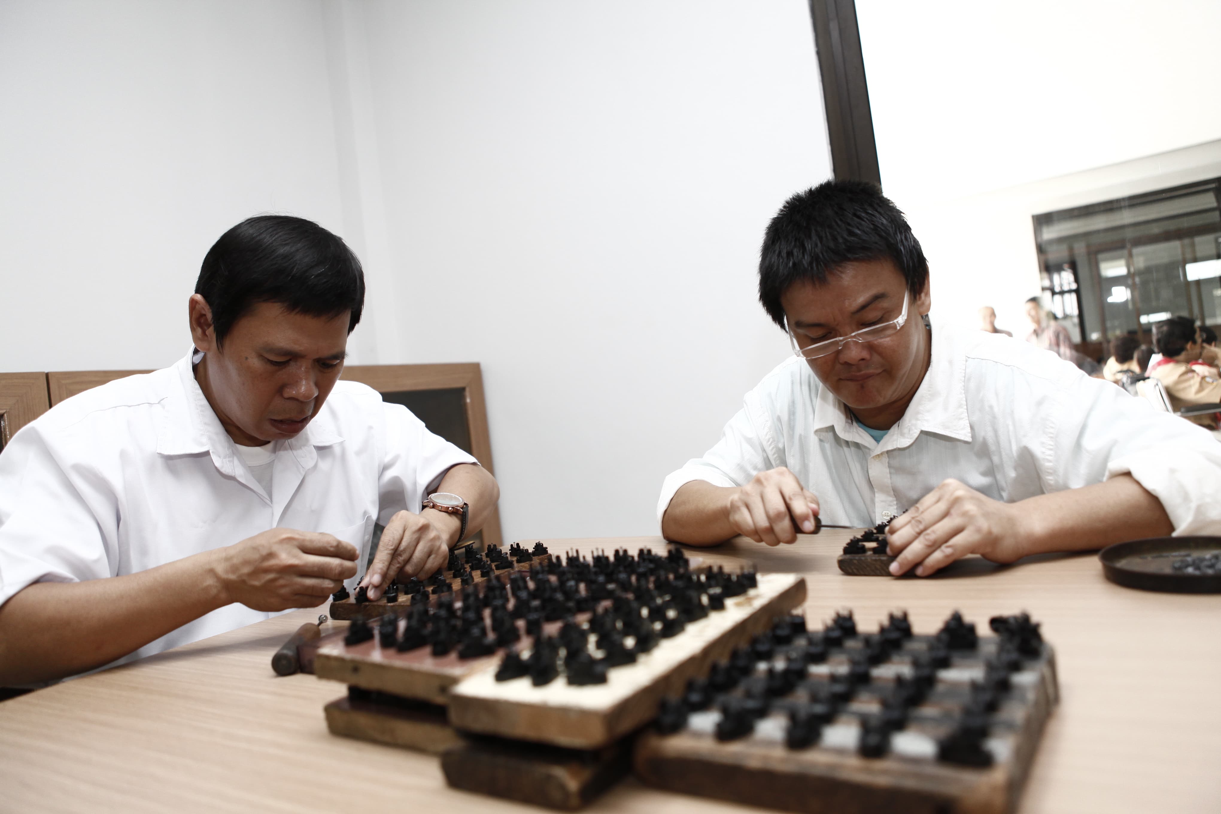 Two workers sit at a wooden table carefully assembling small black components arranged on trays. The setting appears to be an indoor workshop or manufacturing space with neutral walls and bright lighting. Their faces are blurred for privacy, drawing attention to the detailed manual work with the parts in front of them. The overall scene emphasizes concentration, craftsmanship, and hands-on production.