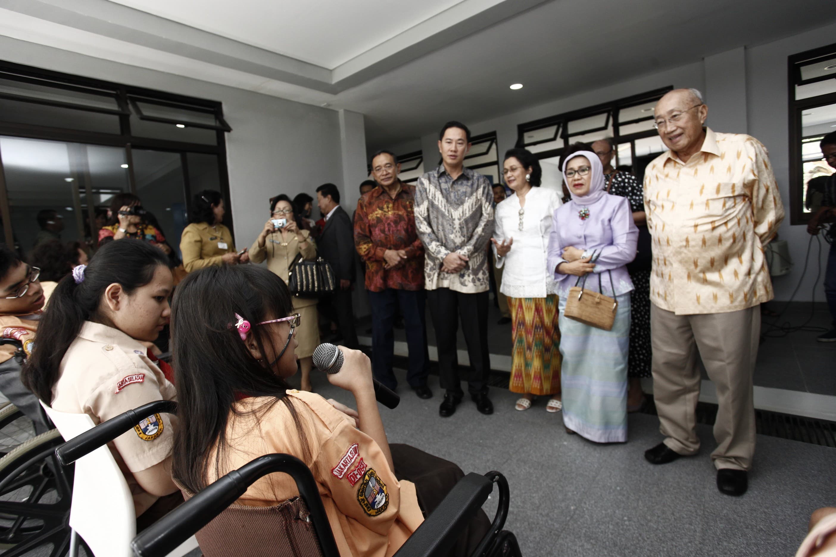 A group of officials and adults stand in front of a classroom area while interacting with seated students. The students wear school uniforms and one girl in a wheelchair speaks into a microphone. Several people in the background observe and take photos, suggesting a formal visit or ceremony. The setting appears to be indoors or in a covered outdoor corridor with large windows and neutral walls.