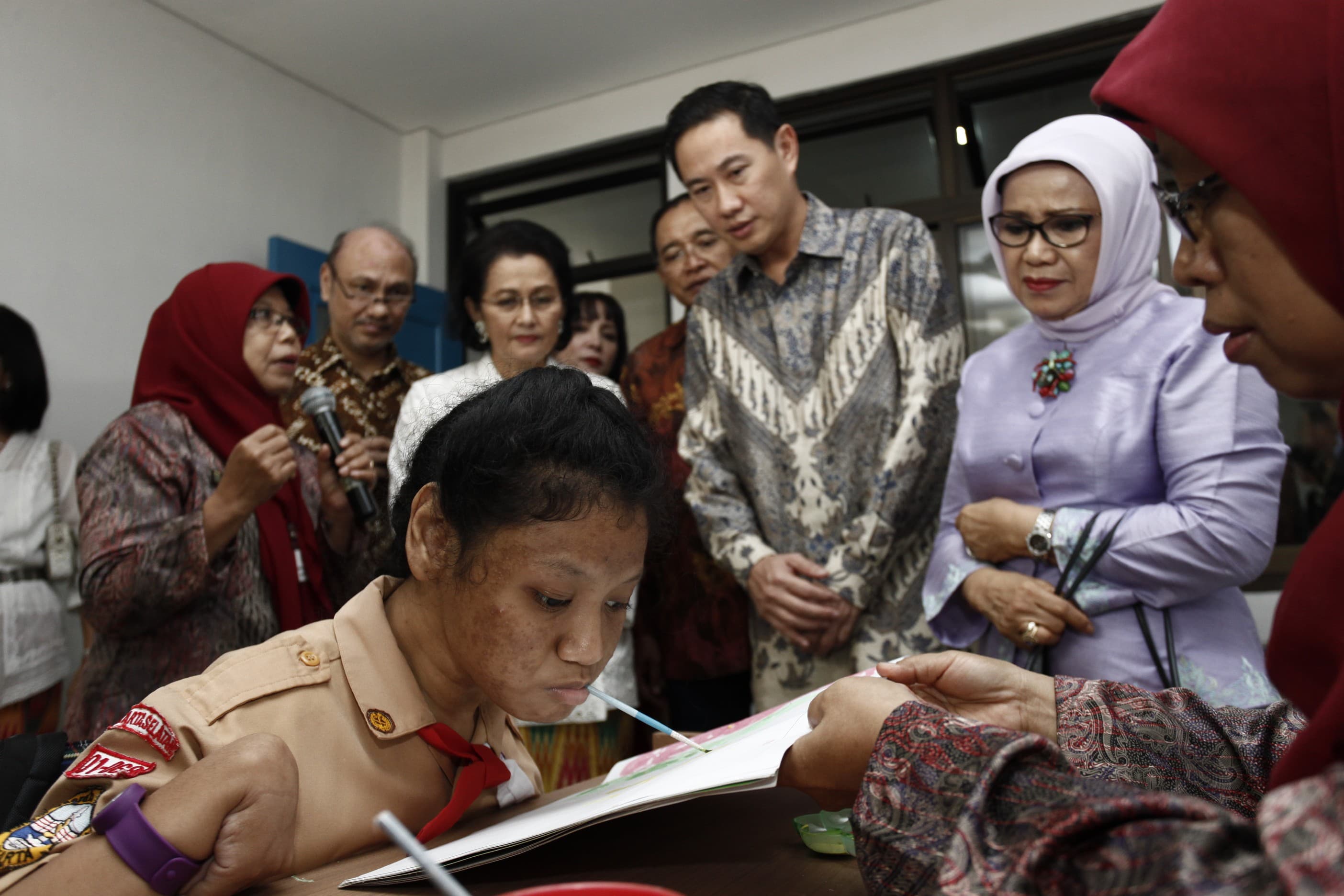 A group of adults gathers around a table in an indoor setting. In the foreground, a person in a scout uniform leans over a document or book while others observe. Several people wear traditional patterned clothing and headscarves, suggesting a formal or cultural event. The atmosphere appears focused and collaborative as they engage with the reading material.