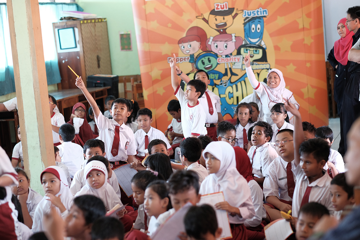 A large group of elementary school children in uniform gather in a classroom for an interactive activity. One boy stands in the middle enthusiastically raising his hand while classmates sit around him holding pencils and notebooks. A colorful educational backdrop with cartoon characters and the names Zul and Justin is visible behind them. The atmosphere appears lively and engaging, suggesting a fun learning or outreach event.