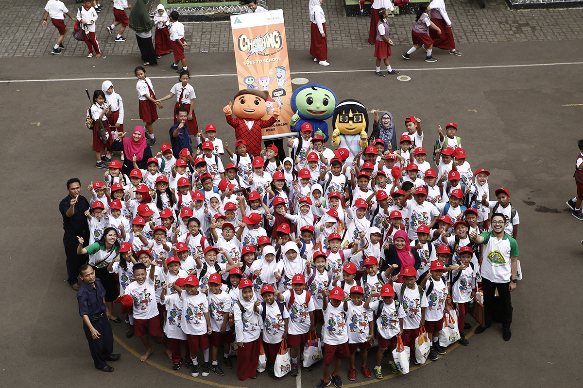 A large group of schoolchildren wearing white shirts and red caps pose together in a school courtyard. Several adults stand around them, and some students in red uniforms are visible in the background. Two people in colorful character costumes and a tall promotional standee are positioned behind the group. The scene suggests a special school event or campaign gathering. The image is taken from an elevated viewpoint, capturing the entire crowd in a circular formation.