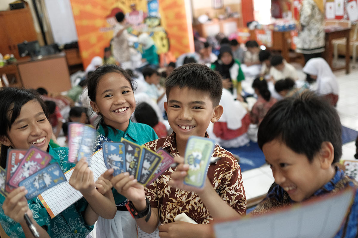 A group of children stand at the front of a classroom holding up colorful cards toward the camera. The classroom is busy, with many other students seated on the floor and working in the background. The children wear patterned shirts and school uniforms, suggesting a school activity or game session. The scene feels lively and playful, with bright colors and a casual learning environment.