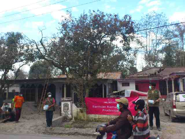 A group of people wearing face masks gather in front of damaged village houses along a roadside. Several individuals appear to be organizing or receiving community relief, with one person holding a box and others standing near a large pink banner. Two people ride past on a motorcycle in the foreground, observing the scene. Trees with sparse foliage and cloudy skies emphasize a somber, post-disaster atmosphere.