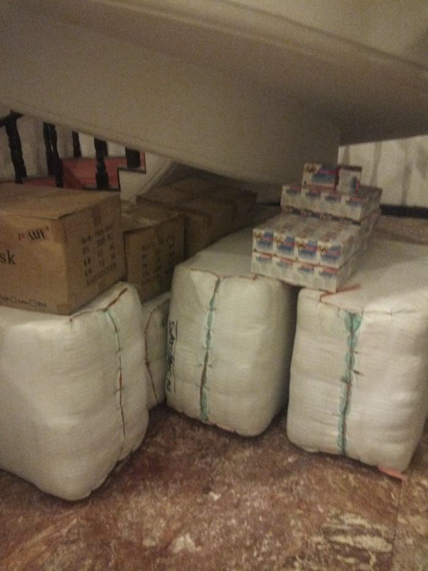 Large white-wrapped bales and cardboard boxes are stacked under an indoor staircase. Smaller cartons are piled on top of the bales, suggesting bulk storage or inventory. The scene appears to be in a residential or small commercial interior with tiled flooring. The overall atmosphere is utilitarian, focused on storage and organization of goods.
