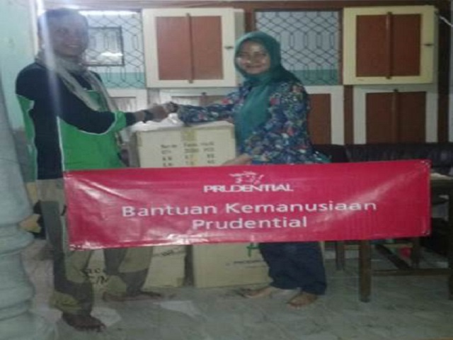 Two adults stand indoors shaking hands in front of stacked cardboard boxes during a donation event. A large red banner with the Prudential name and the Indonesian text "Bantuan Kemanusiaan Prudential" is displayed across the front. The setting appears to be a modest community or residential space with tiled floors and shuttered windows. The scene suggests the handover of humanitarian assistance or relief supplies.