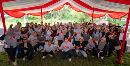 A large group of people pose together under a red and white canopy at an outdoor event. Most participants wear gray shirts, some with red rectangular text on the front, while others are dressed casually. The group smiles, raises hands, and makes cheerful gestures, suggesting a festive or team-building occasion. Green trees and grass surround the tent, adding a natural backdrop to the lively gathering.