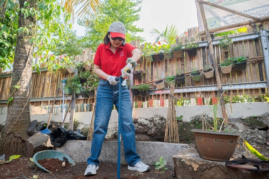 A woman wearing a red shirt, jeans, and a cap works in a garden, using a shovel or digging tool in soil. She is surrounded by potted plants, rocks, and gardening equipment in an outdoor setting. A wooden fence with hanging planters and green foliage forms the background, suggesting a home or community garden. The scene conveys hands-on yard work and care for plants in a bright, natural environment.