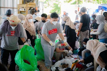 A large group of volunteers work together to sort waste and recyclables in an indoor facility. Many people wear masks, gloves, and casual clothing while handling plastic and other materials. Bright green trash bags and scattered items on the floor emphasize the cleanup activity. The scene suggests a coordinated environmental or community service effort with diverse participants focused on the task.