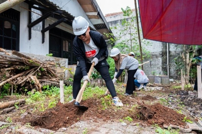 Several volunteers wearing safety helmets are digging and leveling soil in an outdoor area. The scene appears to be near an older building with weathered walls and scattered wooden debris. One person in the foreground uses a shovel while others work in the background, suggesting a community construction or renovation project. The environment includes patches of greenery and a partially covered area with a red fabric canopy.