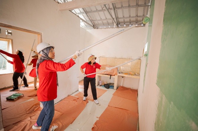 A group of volunteers in red shirts paint the interior walls of a small house. They use long-handled rollers to apply green paint to the walls while standing on a floor covered with protective sheets. The room has an unfinished look with exposed metal roofing and bare window frames. The scene suggests teamwork and community service in a construction or renovation project.