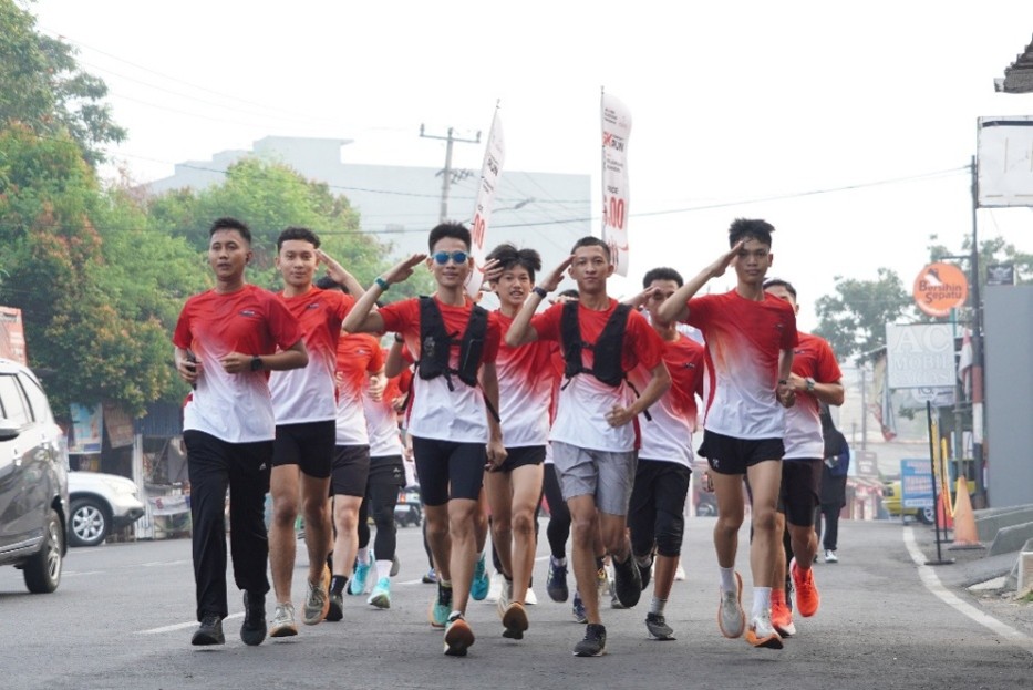 A group of young runners jogs together down a city street. They wear matching red and white gradient athletic shirts and shorts, suggesting a team or organized running event. The background shows urban buildings, trees, and street signs in a hazy morning atmosphere. Some runners carry small backpacks or vests, adding to the sporty and energetic scene.