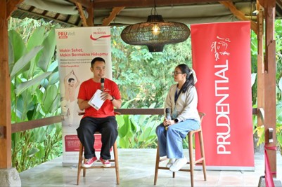 Two speakers sit on stools under a wooden gazebo during an outdoor event, holding microphones as they converse. A large red Prudential banner stands to the right, while a PRUWell Health Indonesia display with text in Indonesian is visible to the left. Lush green foliage surrounds the open pavilion, creating a relaxed and natural atmosphere. The scene suggests an informal health or wellness discussion organized by Prudential.