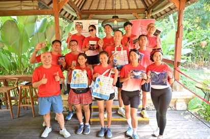 A group of people wearing red shirts pose together on a wooden deck under a thatched roof structure. Several individuals hold up printed materials, tote bags, and small items toward the camera. Lush green tropical plants surround the open-air setting, suggesting an outdoor event or gathering. The group smiles and gestures enthusiastically, creating a cheerful and energetic atmosphere.