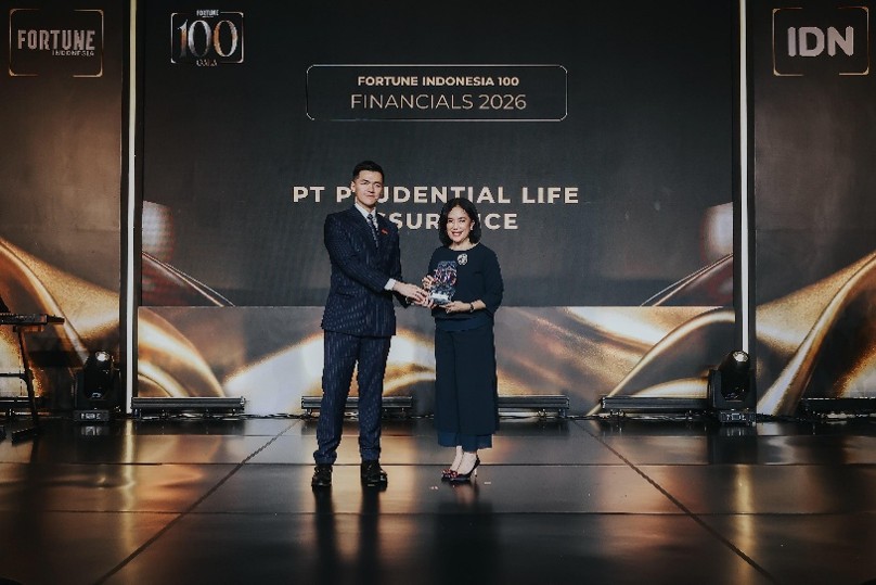Two business professionals stand on stage during an award presentation at a Fortune Indonesia 100 Financials 2026 event. The man in a suit hands a trophy or plaque to a woman dressed in dark attire, both posing for a photo. The backdrop displays the text "FORTUNE INDONESIA 100 FINANCIALS 2026" and "PT PRUDENTIAL LIFE INSURANCE" along with decorative golden abstract visuals. Stage lighting and side panels with "FORTUNE" and "IDN" text frame the central scene.