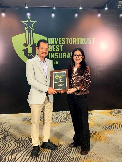 Two business professionals stand together holding an award plaque in front of a large event backdrop. The backdrop prominently displays the text 'INVESTORTRUST BEST INSURANZ 2026' with a stylized shield and star graphic. The man on the left wears a light-colored blazer and trousers, while the woman on the right wears dark slacks and a patterned blouse. The setting appears to be an indoor awards ceremony or corporate recognition event with patterned carpet and stage lighting.