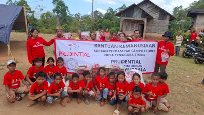 A group of children and adults pose together outdoors in front of a large humanitarian aid banner. The banner text mentions "Bantuan Kemanusiaan" and support for earthquake victims in Flores, Nusa Tenggara Timur. Many participants wear matching red shirts, and simple wooden houses and trees are visible in the background, indicating a rural village setting. The scene conveys solidarity, relief support, and community spirit.