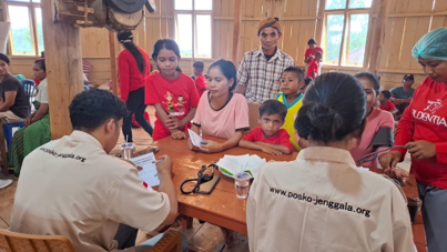 A group of adults and children gather around a wooden table in a village hall for a community outreach or health registration activity. Two staff members sit at the table, writing on forms and speaking with the people waiting in line. The room has wooden walls, large windows, and simple furnishings, creating a warm, informal setting. Text on the back of one staff member’s shirt reads "www.posko-jenggala.org," indicating an organized relief or support effort.