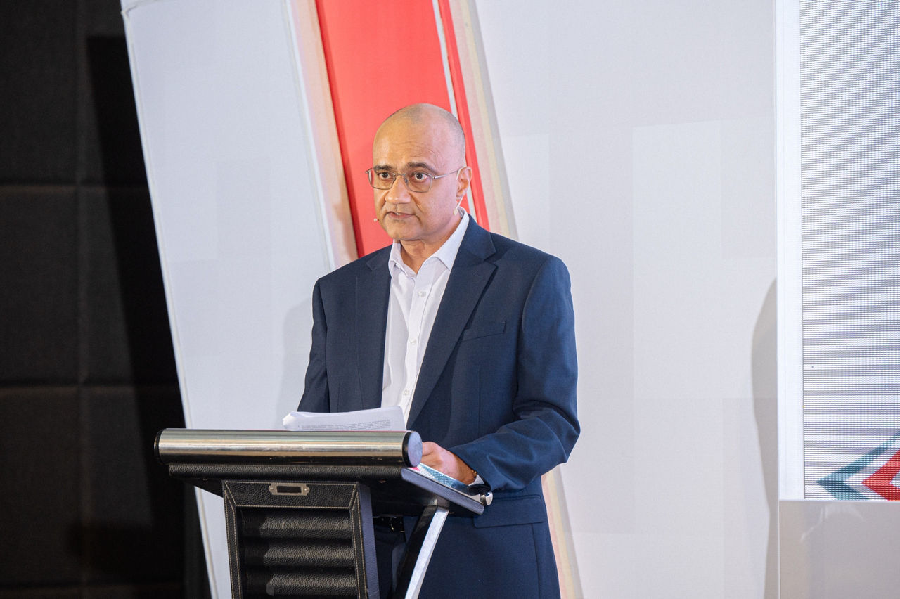 A formally dressed person stands behind a lectern delivering a speech. The individual wears a dark suit and white shirt while holding papers and a tablet or notebook. The background features large vertical panels in white and red tones, suggesting a professional conference or corporate event. Stage lighting highlights the speaker and podium, creating a focused presentation setting.