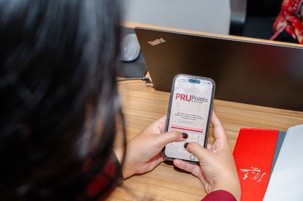 A person holds a smartphone displaying the PRUPoints interface while seated at a wooden desk. A laptop is open in the background, suggesting a work or office environment. The screen shows text related to PRUPoints with a clean, white layout and red accents. Nearby, a red notebook or folder adds a pop of color to the scene.