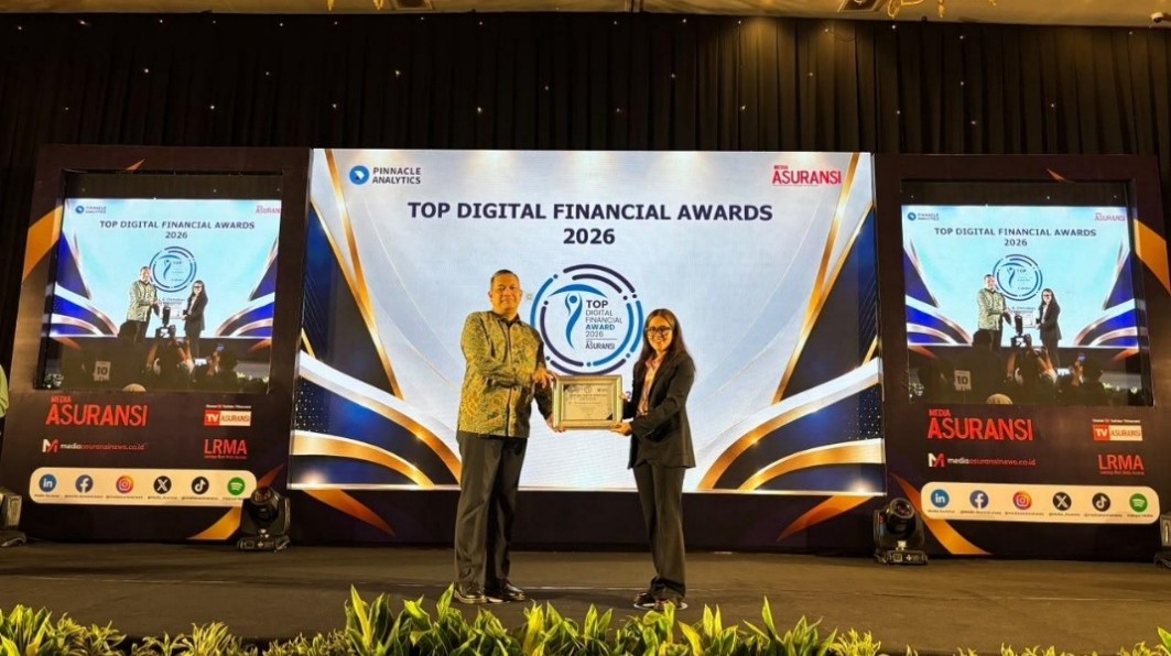 Two people stand on a stage during the Top Digital Financial Awards 2026 event. One person presents a trophy while the other holds a framed certificate in front of a large illuminated backdrop. The main screen displays the award title and circular emblem, flanked by two side screens with event graphics and sponsor names including Asuransi and LRMA. Decorative lighting and plants line the front of the stage, creating a formal and celebratory atmosphere.