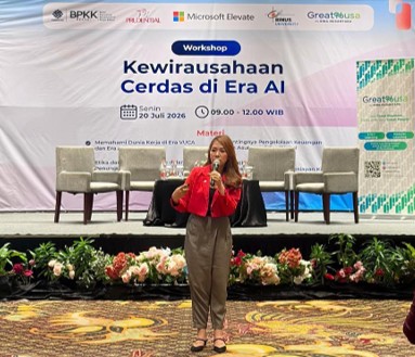 A woman stands on a decorated stage speaking at a workshop titled "Kewirausahaan Cerdas di Era AI." The event backdrop shows the date Senin 20 Juli 2026 and the time 09.00 - 12.00 WIB, along with several organizer names and partner graphics. Behind her are chairs and a table arranged for a panel discussion, while the foreground features floral arrangements on a patterned carpet. The atmosphere suggests a formal business and technology event focused on smart entrepreneurship in the age of artificial intelligence.