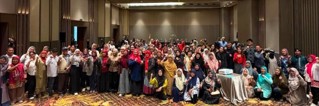 A large group of people pose together in a spacious hotel ballroom. The attendees stand and sit in several rows, many smiling and gesturing toward the camera. The group includes adults of diverse backgrounds wearing a variety of colorful outfits, including headscarves and casual clothing. The room features tall walls, warm lighting, and patterned carpet, suggesting a formal event or conference setting.