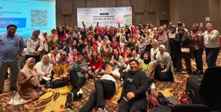 A large group of people pose together in a conference room after an event or seminar. Participants of various ages stand and sit closely, smiling at the camera, with some seated on the patterned carpet in front. A presentation screen with a QR code and a banner at the back indicate an organized workshop or training session. The atmosphere appears friendly and celebratory, suggesting the conclusion of a group activity.