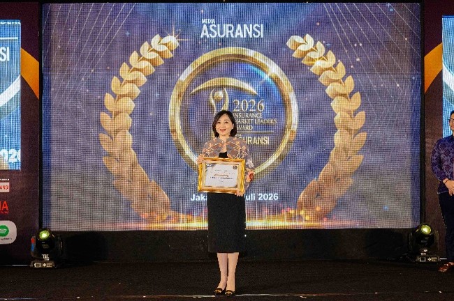 A woman stands on a stage holding a framed certificate during an insurance industry award ceremony. Behind her, a large digital screen displays a golden laurel wreath and the year 2026 prominently. The backdrop includes the text "ASURANSI" and other event details, suggesting a formal recognition event in Jakarta. The lighting and decor create a celebratory and professional atmosphere.