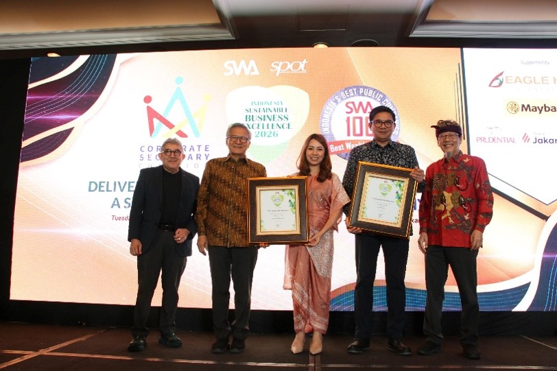 Five adults stand on a stage during a corporate award ceremony, with two individuals holding large framed certificates. The backdrop screen displays event graphics and text including "Corporate Secretary", "SWA", "spot", and "SWA 100". The setting appears to be an indoor conference or business event with formal and semi-formal attire. The overall atmosphere is professional and celebratory, highlighting recognition and achievement.