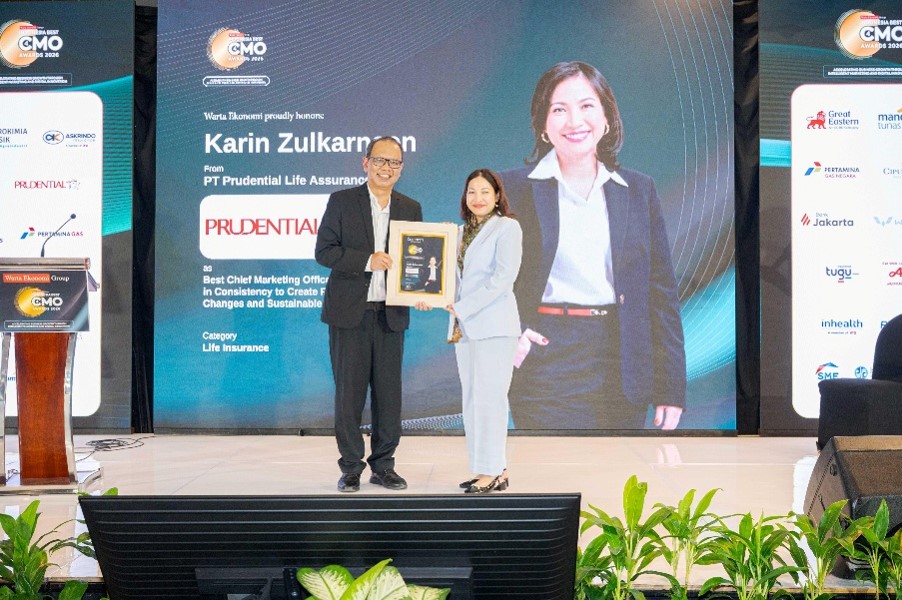 Two business professionals stand on a stage during a formal award ceremony, holding a framed plaque together. Behind them, a large screen displays the name Karin Zulkarnaen and PT Prudential Life Assurance, along with event and category details related to life insurance. The setting is a brightly lit conference hall with banners, sponsor panels, and green plants along the front of the stage. The overall atmosphere is professional and celebratory, highlighting corporate recognition and achievement.