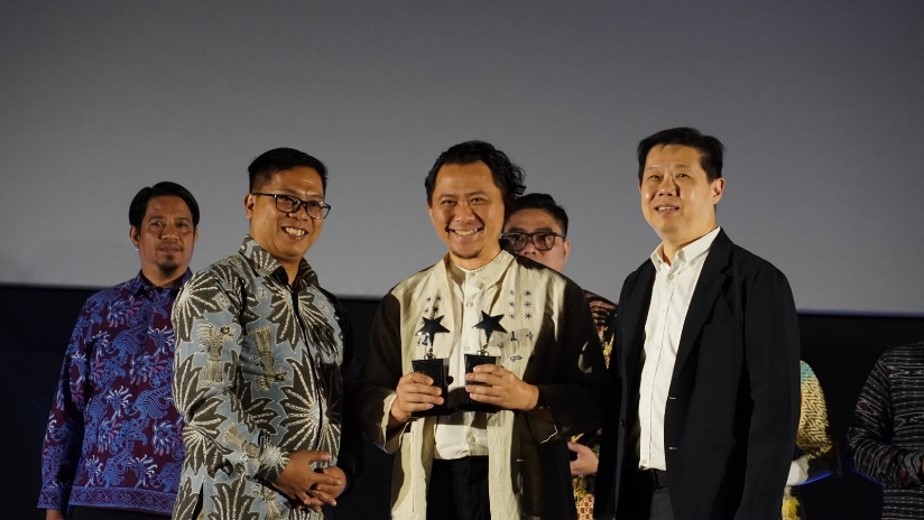 A man at the center of the image smiles while holding two star-shaped trophies. He is surrounded by four other men standing on a stage in front of a large screen. Several of the men wear patterned batik shirts, while others are dressed in suits, suggesting a formal or semi-formal event. The scene appears to capture a celebratory moment during an award ceremony.
